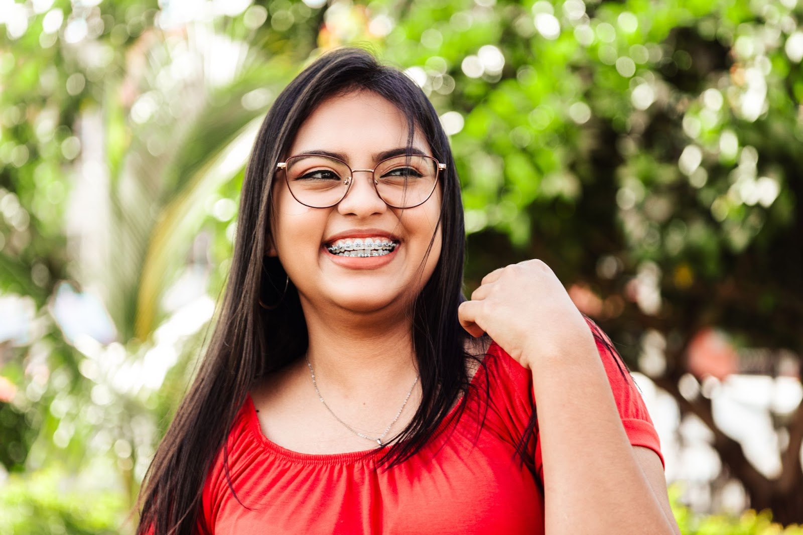 Smiling young woman with braces in a red shirt, outdoors with greenery, representing orthodontic care and patient confidence.